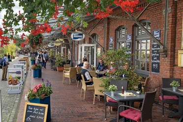 Menschen sitzen vor Backsteinfassaden im Schaufenster Fischereihafen Bremerhaven bei Speisen und Getränken.