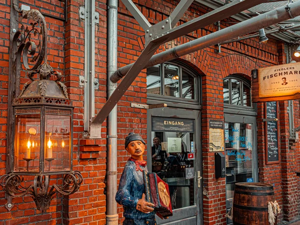 Schaufenster Fischereihafen DSC00743 (c) Birgit Hass_Erlebnis Bremerhaven.jpg Backsteinfassade mit großer Laterne, Holzfigur eines Akkordeonspielers und Eingang zu Fiedlers Fischmarkt.