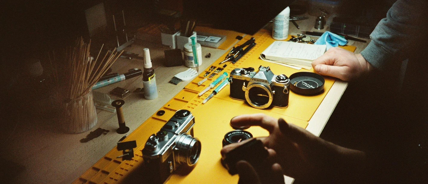 Cologne-Camera-Service-Analog-Technik-Foto Two analog cameras and several lenses on a table, accompanied by hands waiting for equipment.