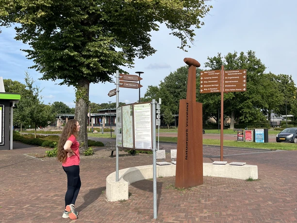 Das Symbol für den Naturpark Bourtanger Moor - Veenland - der Oplegger..jpeg Frau betrachtet Infotafel mit Wegweisern und rostfarbener Skulptur im Naturpark Bourtanger Moor.