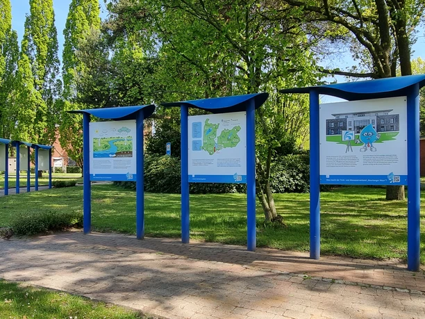 Trink-Wasser-Erlebnis-Pfad Geeste 1.jpg Reihe blauer Informationstafeln mit Illustrationen und Texten in einer grünen Parklandschaft.