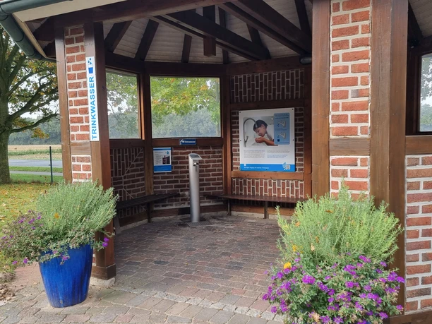 Trink-Wasser-Erlebnis-Pfad Geeste 3.jpg Backsteingebäude mit überdachter Trinkwasserstelle, Infotafeln und Blumenbeeten am Wegesrand.