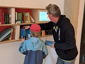 Ausstellung Romantik Bad Rehburg Kind und Erwachsener vor einem Bücherregal in der Ausstellung Romantik Bad RehburgChild and adult in front of a bookshelf in the Romantik Bad Rehburg exhibitionBarn og voksen foran en bogreol i udstillingen Romantik Bad RehburgKind en volwassene voor een boekenplank in de tentoonstelling Romantik Bad Rehburg