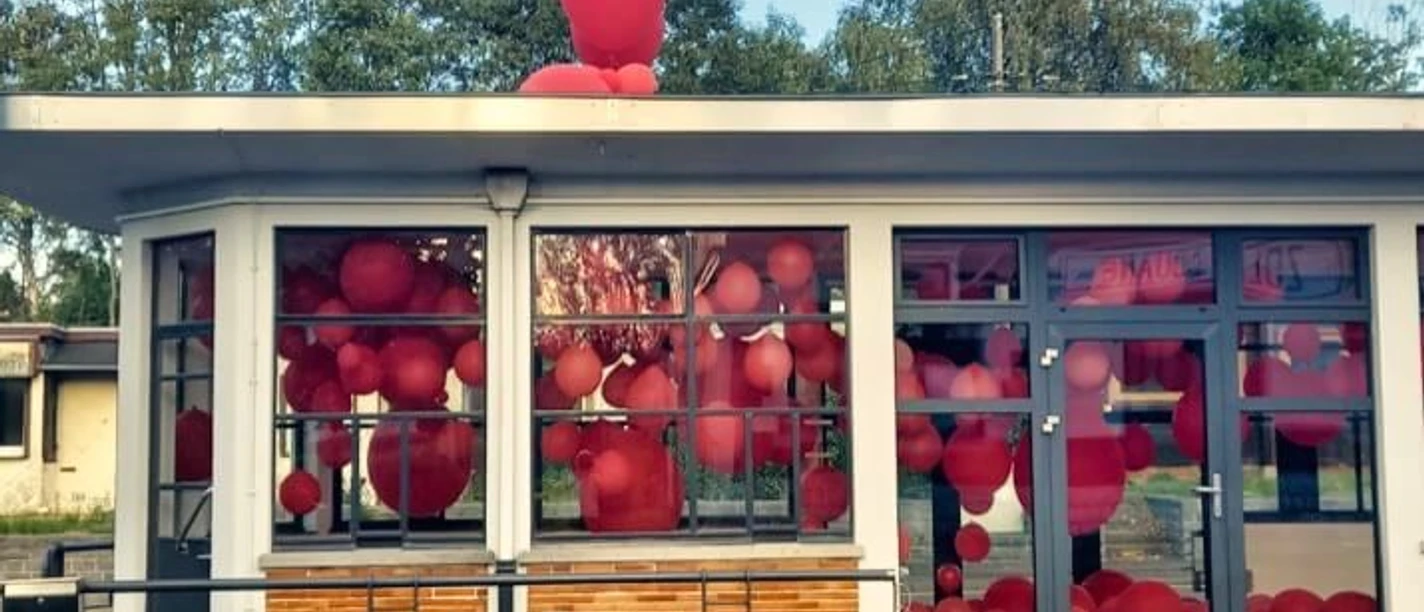 Ein Gebäude mit gläsernem Eingangsbereich, gefüllt mit zahlreichen roten Ballons, vor einer grünen Baumkulisse.