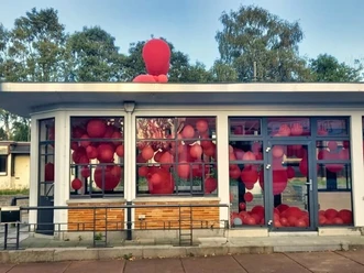 20 Jahre KuK 1 (c) KuKuK e.V..jpg Ein Gebäude mit gläsernem Eingangsbereich, gefüllt mit zahlreichen roten Ballons, vor einer grünen Baumkulisse.