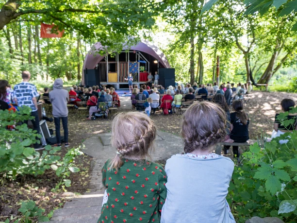 Zwei Kinder mit geflochtenem Haar sitzen im Wald und blicken auf eine Bühne vor Publikum.