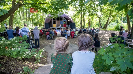 Waldbühne 2 (c) KuKuK e.V..jpg Zwei Kinder mit geflochtenem Haar sitzen im Wald und blicken auf eine Bühne vor Publikum.
