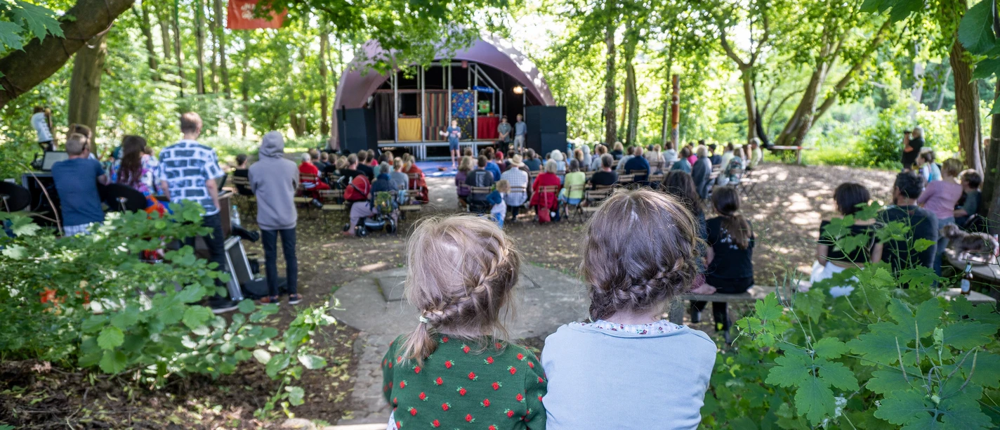 Waldbühne 2 (c) KuKuK e.V..jpg Zwei Kinder mit geflochtenem Haar sitzen im Wald und blicken auf eine Bühne vor Publikum.
