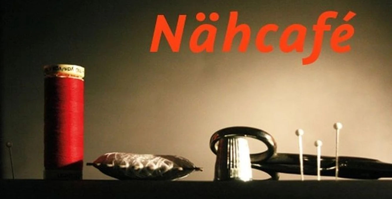 Nähcafe in der Kombüse Oberndorf Nahaufnahme mit rotem Garn, Nadelkissen und Fingerhut unter warmem Licht, Schriftzug Nähcafé