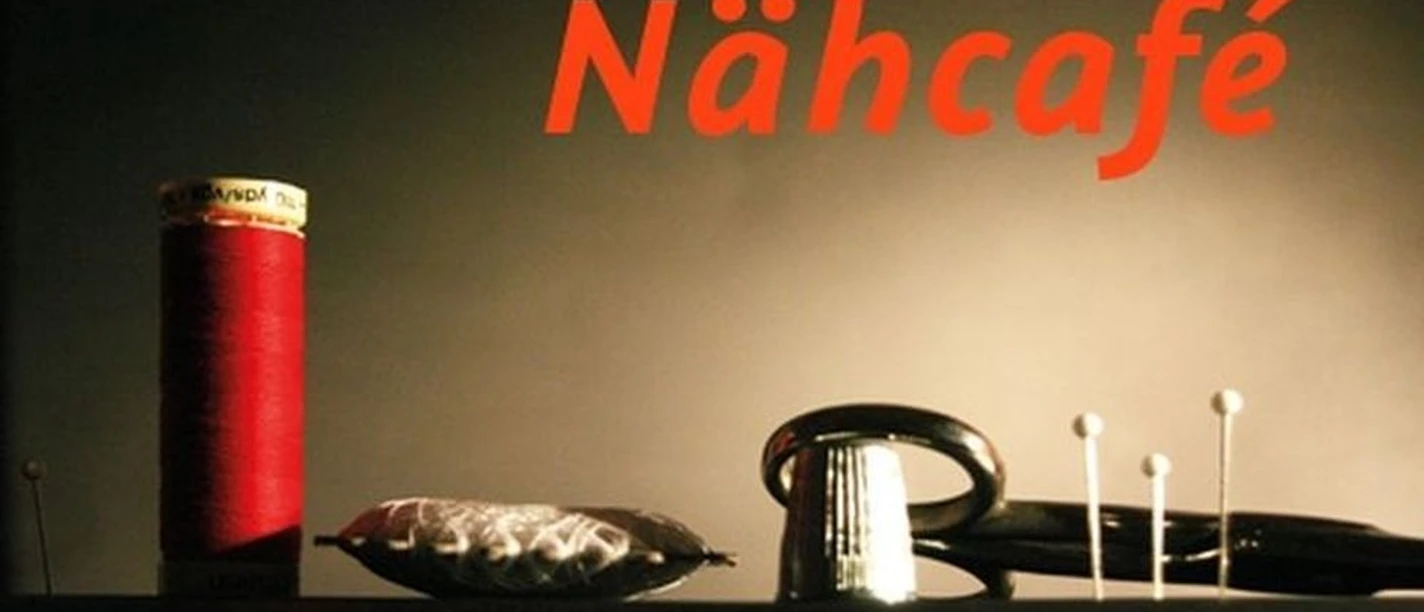 Nähcafe in der Kombüse Oberndorf Nahaufnahme mit rotem Garn, Nadelkissen und Fingerhut unter warmem Licht, Schriftzug Nähcafé