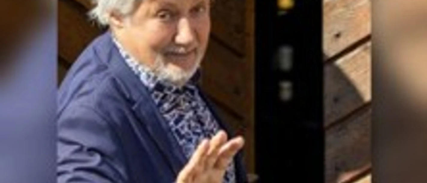 Ein Mann mit grau-meliertem Haar in einem blauen Sakko hebt lächelnd die Hand zum Gruß.