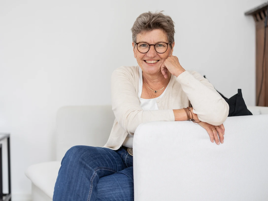 Foto_Bagge2.jpg Ältere Frau mit Brille sitzt auf Sofa und lächelt freundlich in heller Wohnung.