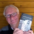 Foto_Johannes Bollen.jpg Ein älterer Mann hält ein Buch und lächelt freundlich vor einer Holzwand.