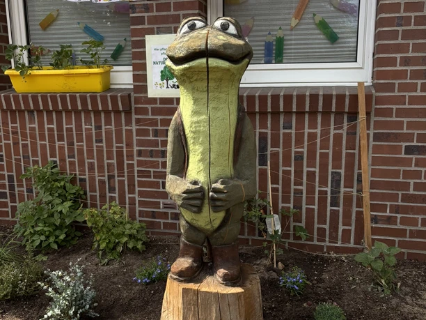 MOORex ist das Maskottchen der Naturpark-Schulen.JPEG Holzfigur eines freundlich lächelnden Froschs in grüner Farbe vor einem Schulgebäude mit Blumenbeet