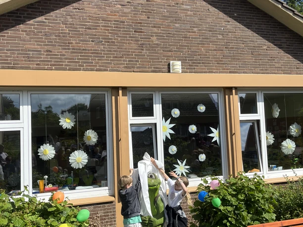 Feierlich wird MOORex enthüllt.jpeg Zwei Kinder enthüllen im Garten einer Schule eine grüne Moorfigur vor geschmückten Fenstern.