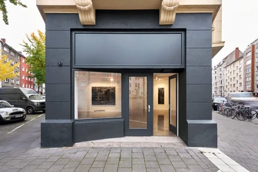 Installationsansicht von Laurie Smith, Brick boys, bei Gathering Cologne Moderner Galerieeingang mit gläserner Fassade und Kunstwerken im Inneren in urbaner Umgebung.