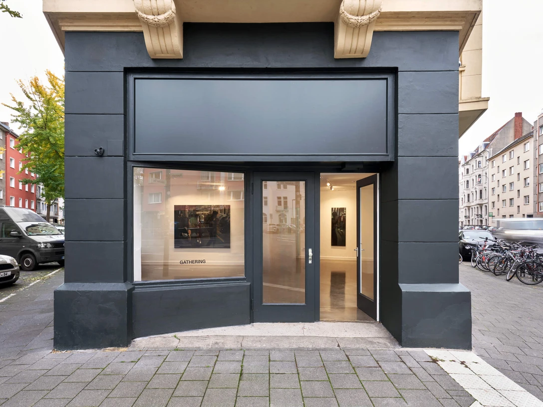 Installationsansicht von Laurie Smith, Brick boys, bei Gathering Cologne Moderner Galerieeingang mit gläserner Fassade und Kunstwerken im Inneren in urbaner Umgebung.Modern gallery entrance with glass façade and works of art inside in an urban setting.