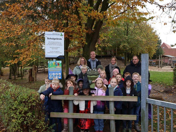 Auf Augenhöhe mit MOORex.JPG Kinder und Erwachsene stehen lachend im Herbstlaub neben einer Froschfigur auf einem Schulhof.