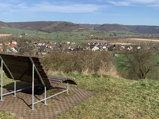 Von einer Liegebank aus genießt man den Ausblick auf das im Tal gelegene Amelunxen, im Hintergrund die Höhen des Weserberglands.