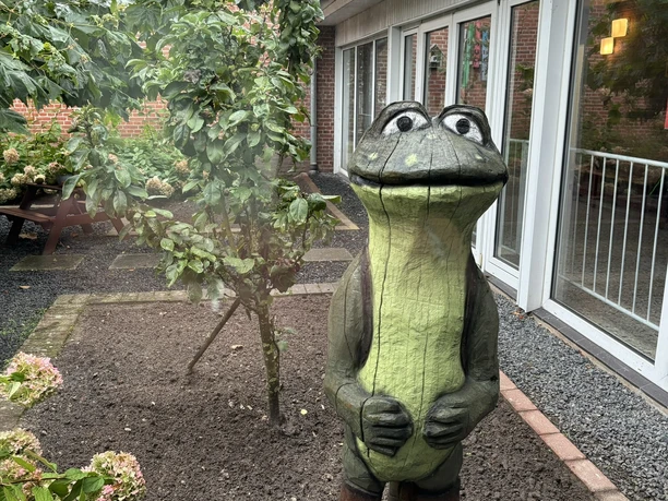 Willkommen MOORex in der Grundschule Adorf.JPEG Holzfigur eines freundlich lächelnden Froschs mit Kleidung im Schulgarten vor roten Backsteingebäuden