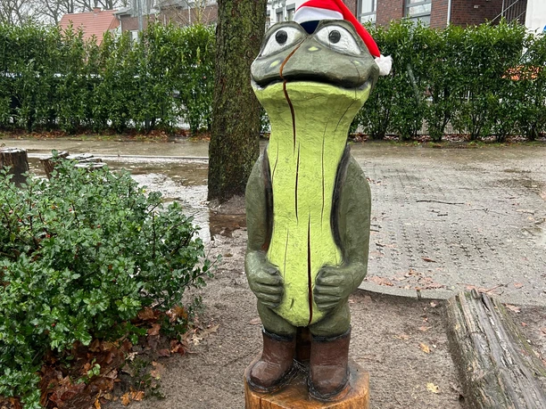 Moorex.JPG Holzfigur eines Frosches mit roter Weihnachtsmütze und braunen Stiefeln auf nassem Platz.