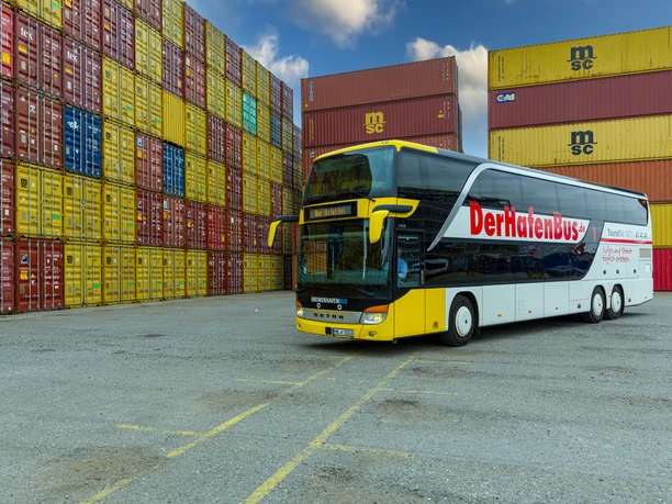 HafenBus S7345 (c) Helmut Gross_Erlebnis Bremerhaven_bearbeitet mit KI.jpg Ein gelb-weißer Doppelstockbus steht vor bunten, gestapelten Containern im Hafen.