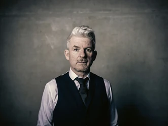 Heinz Strunk25-3©Dennis Dirksen-web.jpg Mann mit grauen Haaren in Weste und Hemd blickt ernst in die Kamera vor schlichtem Hintergrund.