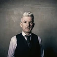 Heinz Strunk25-3©Dennis Dirksen-web.jpg Mann mit grauen Haaren in Weste und Hemd blickt ernst in die Kamera vor schlichtem Hintergrund.