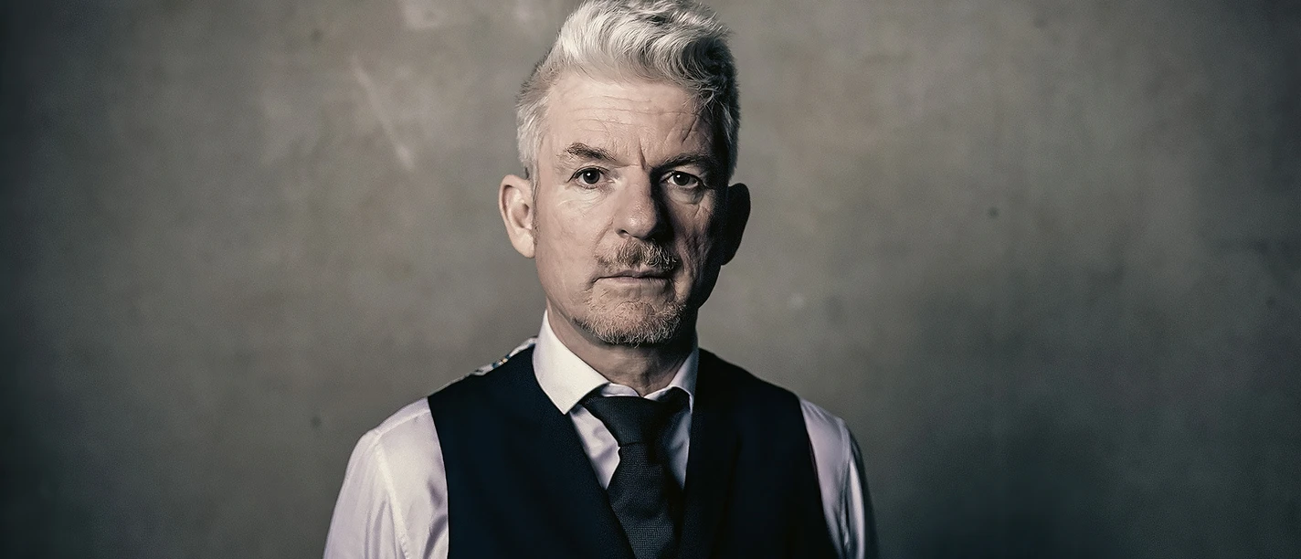 Heinz Strunk25-3©Dennis Dirksen-web.jpg Mann mit grauen Haaren in Weste und Hemd blickt ernst in die Kamera vor schlichtem Hintergrund.