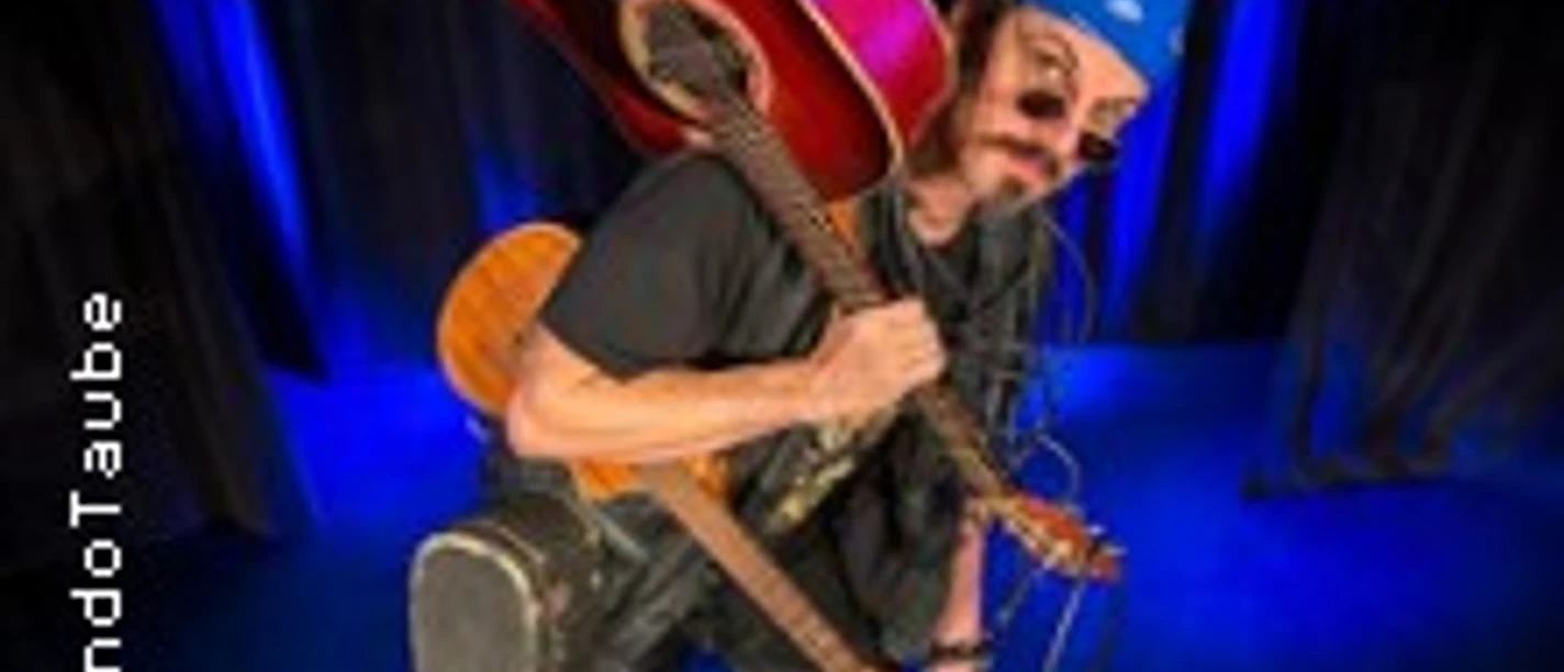 Mann mit blauer Bandana und Sonnenbrille trägt zwei Gitarren und einen bunten Koffer auf der Bühne.