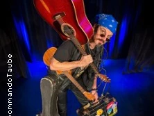 Mann mit blauer Bandana und Sonnenbrille trägt zwei Gitarren und einen bunten Koffer auf der Bühne.