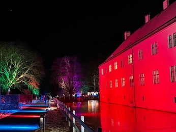 Farbig beleuchtetes Schloss Benkhausen bei Nacht, mit Lichtinstallationen im Garten und Teichreflexion.