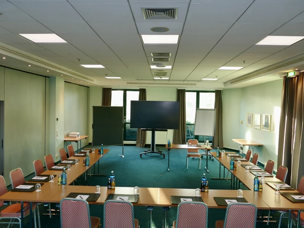 Hotel Amadeus Frankfurt - Tagungsraum U-Form Bestuhlung Tagungsraum in U-Form Bestuhlung mit eingedeckten Tagungsgetränken und Blick auf eine Präsentationsfläche.