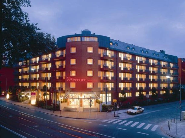 Mercure Hotel & Residenz Frankfurt Messe Außenansicht eines beleuchteten Hotelgebäudes in der Dämmerung.