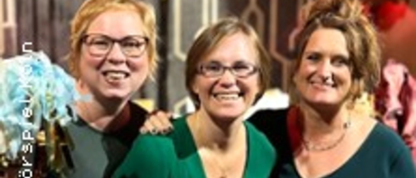 Drei lächelnde Personen in festlicher, farbenfroher Umgebung posieren zusammen vor geometrischer Tapete.