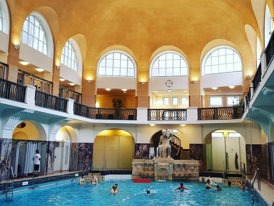 Schwimmhalle im Jugendstil mit hohen Decken und Torbögen. Sie ist warm beleuchtet und im Becken schwimmen einige Personen entspannt.