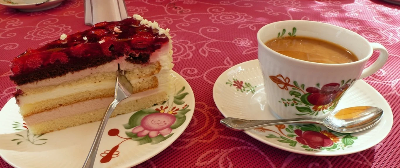 Kaffee und Kuchen Tasse Kaffee mit Milch und Stück Himbeertorte auf floralem Porzellan vor roter Tischdecke