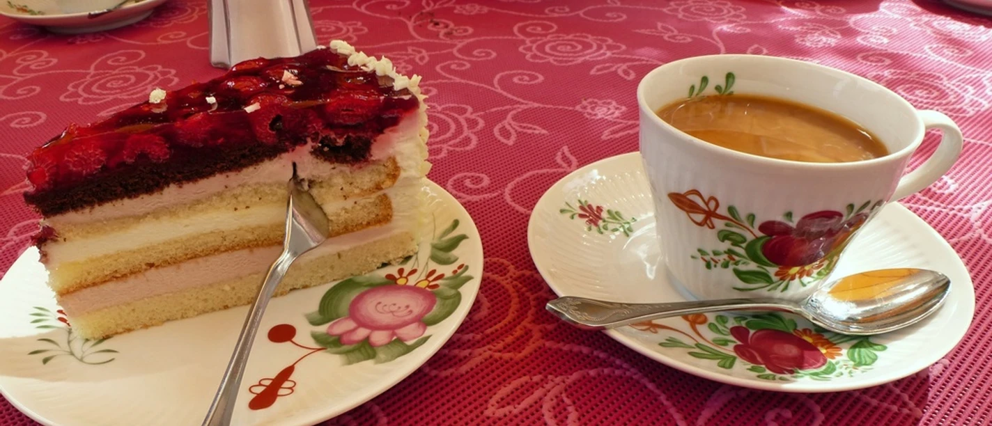 Kaffee und Kuchen Tasse Kaffee mit Milch und Stück Himbeertorte auf floralem Porzellan vor roter Tischdecke