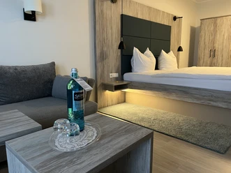 Im Hotel Grasberger Hof Modernes Hotelzimmer mit Doppelbett, grauer Couch, Holzmöbeln und sanfter Beleuchtung.