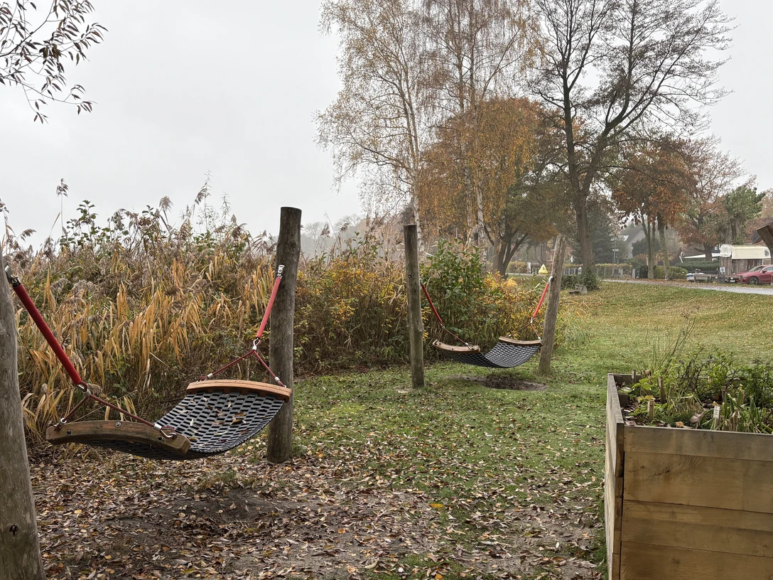 Rastplatz vor dem Naturparkhaus.JPG Holzpfosten mit Netzschaukeln auf einer Wiese vor herbstlicher Vegetation und Häusern im Hintergrund.