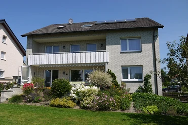Ferienwohnung Iburgblick Zweistöckiges Haus mit hellem Klinker, Balkon und blühendem Garten bei sonnigem Wetter.