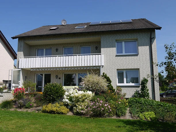 Ferienwohnung Iburgblick Zweistöckiges Haus mit hellem Klinker, Balkon und blühendem Garten bei sonnigem Wetter.