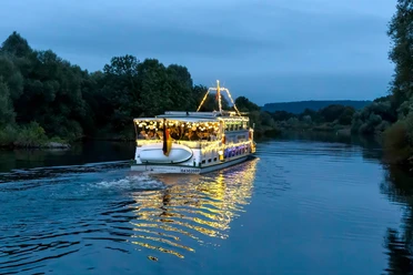 Lampionfahrt, Weiße Flotte Mülheim Ein beleuchtetes Schiff gleitet sanft über einen Fluss, umgeben von Bäumen am Abendhimmel.