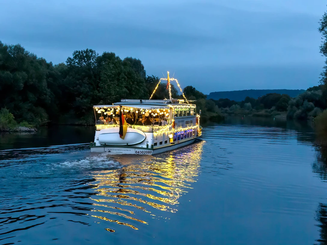 Lampionfahrt, Weiße Flotte Mülheim Ein beleuchtetes Schiff gleitet sanft über einen Fluss, umgeben von Bäumen am Abendhimmel.