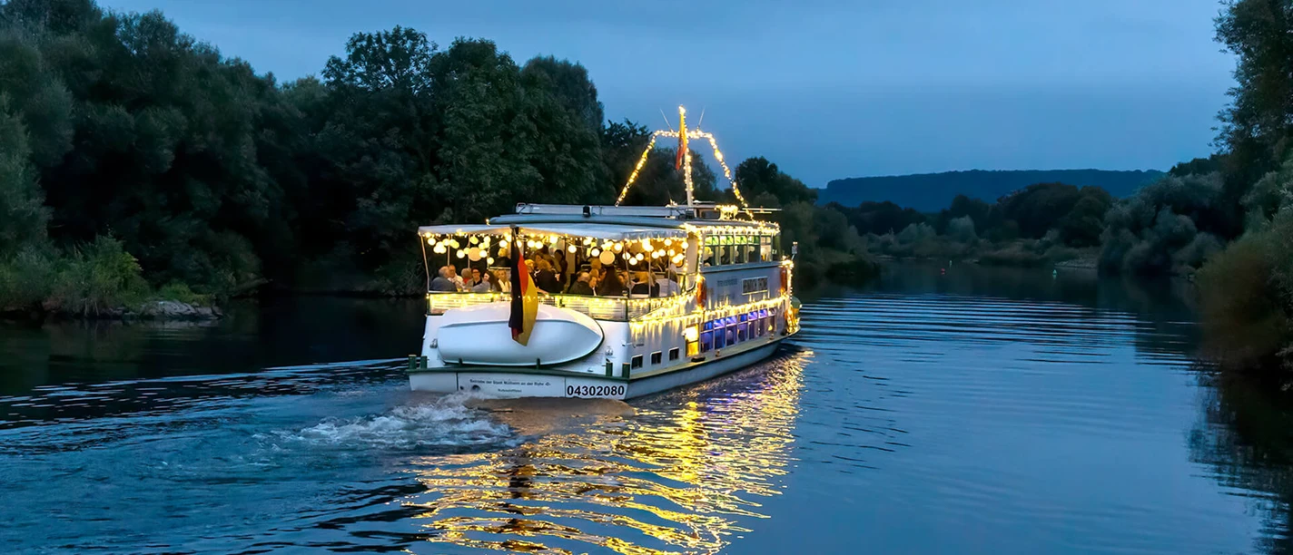 Lampionfahrt, Weiße Flotte Mülheim Ein beleuchtetes Schiff gleitet sanft über einen Fluss, umgeben von Bäumen am Abendhimmel.