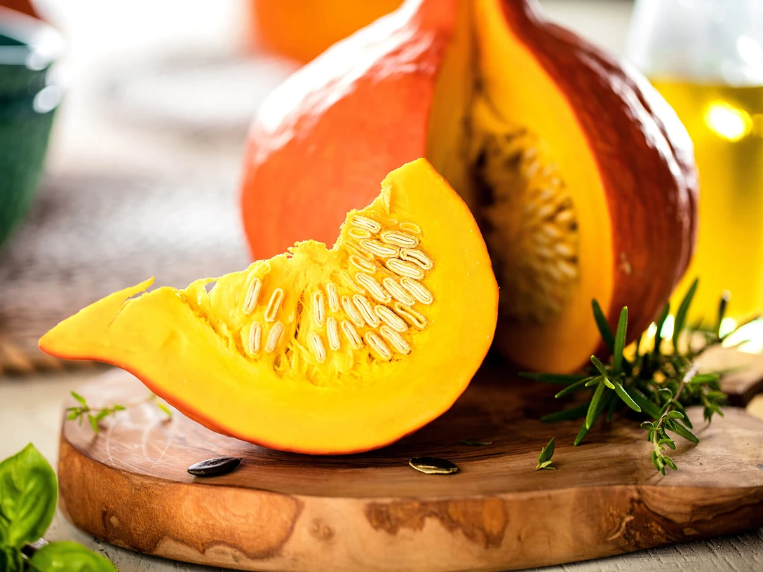 hokkaido-kuerbis-holzbrett.jpg Ein aufgeschnittener Hokkaido-Kürbis mit Kernen auf einem Holzbrett, umgeben von Kräutern.