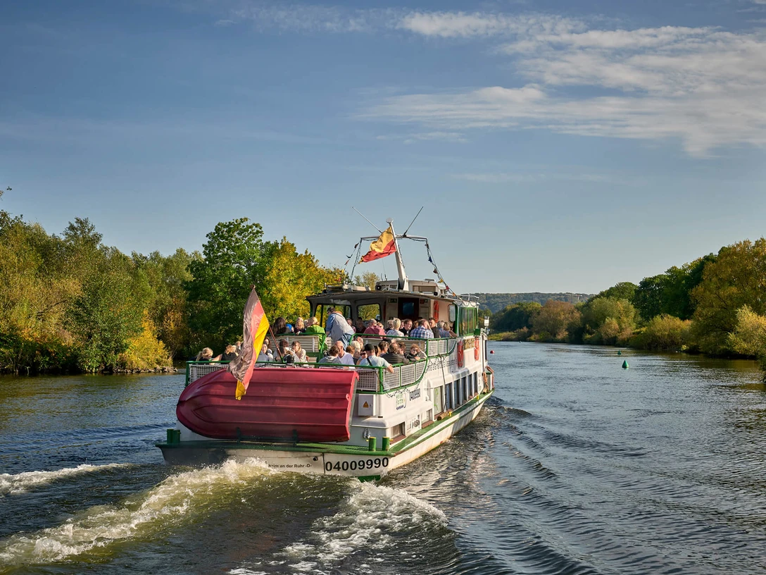 fahrgast-freideck-linienfahrt-weisse-flotte-sommer-ruhr.jpg Ein Ausflugsschiff fährt auf einem Fluss, umgeben von grüner Natur an einem sonnigen Tag.