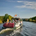 fahrgast-freideck-linienfahrt-weisse-flotte-sommer-ruhr.jpg Ein Ausflugsschiff fährt auf einem Fluss, umgeben von grüner Natur an einem sonnigen Tag.