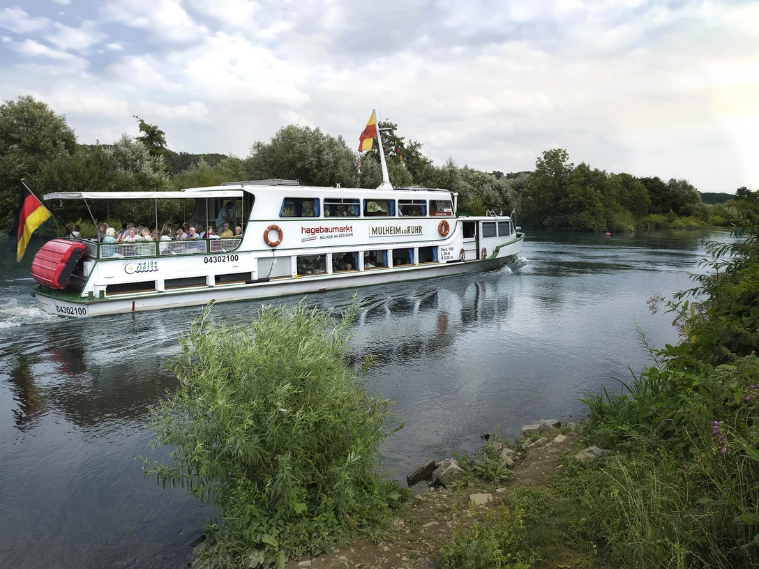 Weiße Flotte, Mülheim an der Ruhr Ein Ausflugsschiff fährt auf einem ruhigen Fluss, umgeben von grünen Bäumen.