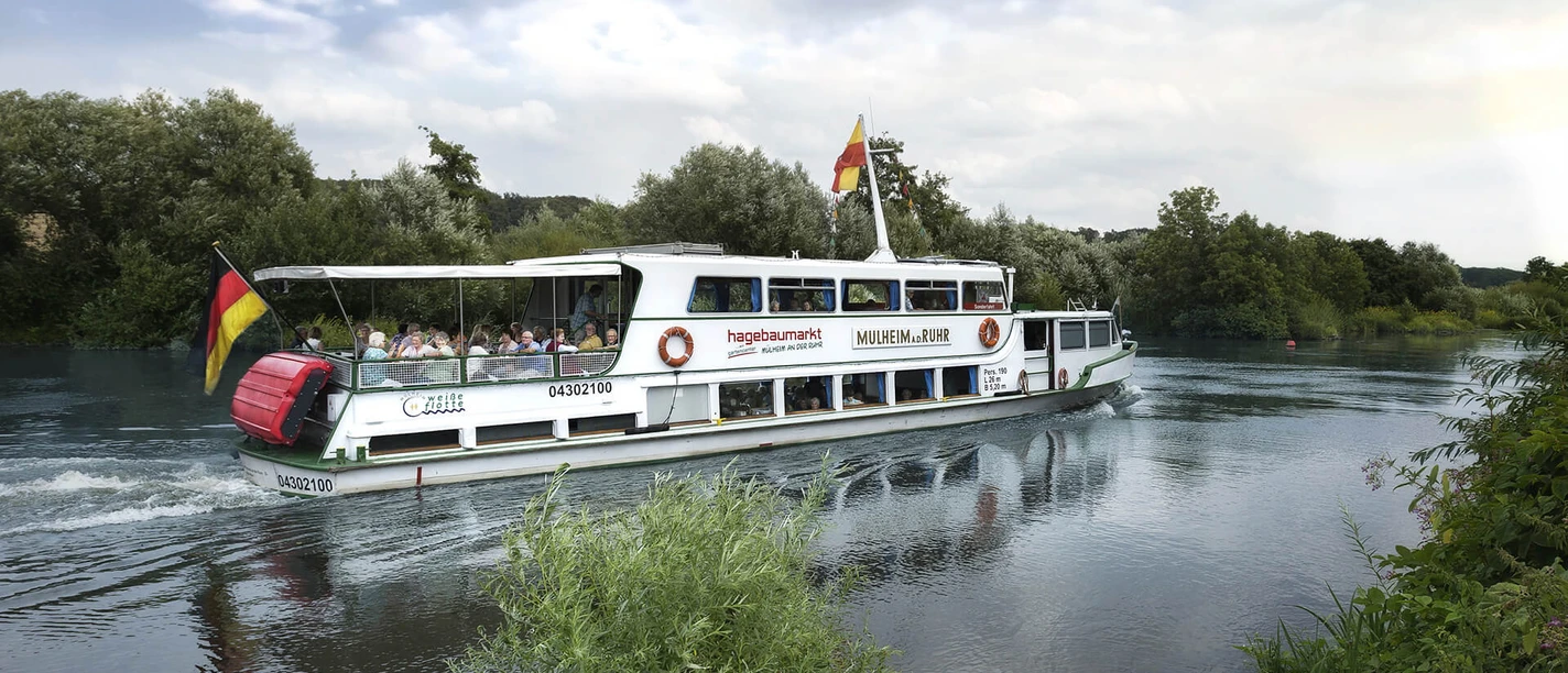 Weiße Flotte, Mülheim an der Ruhr Ein Ausflugsschiff fährt auf einem ruhigen Fluss, umgeben von grünen Bäumen.